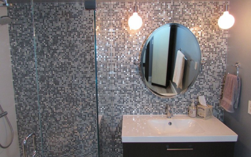 Grande espelho acima da pia do banheiro, com azulejos cinza, bege e preto atrás da parede